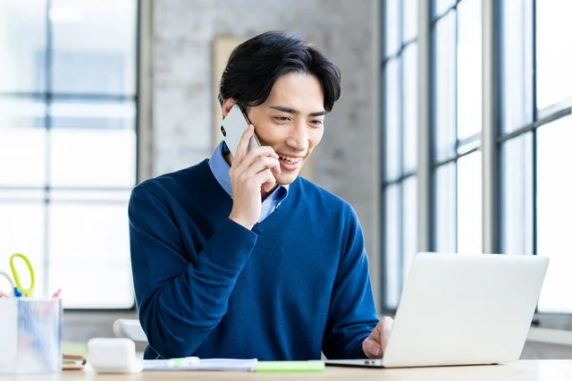 スマートフォンで通話しながらノートパソコンに向かう男性