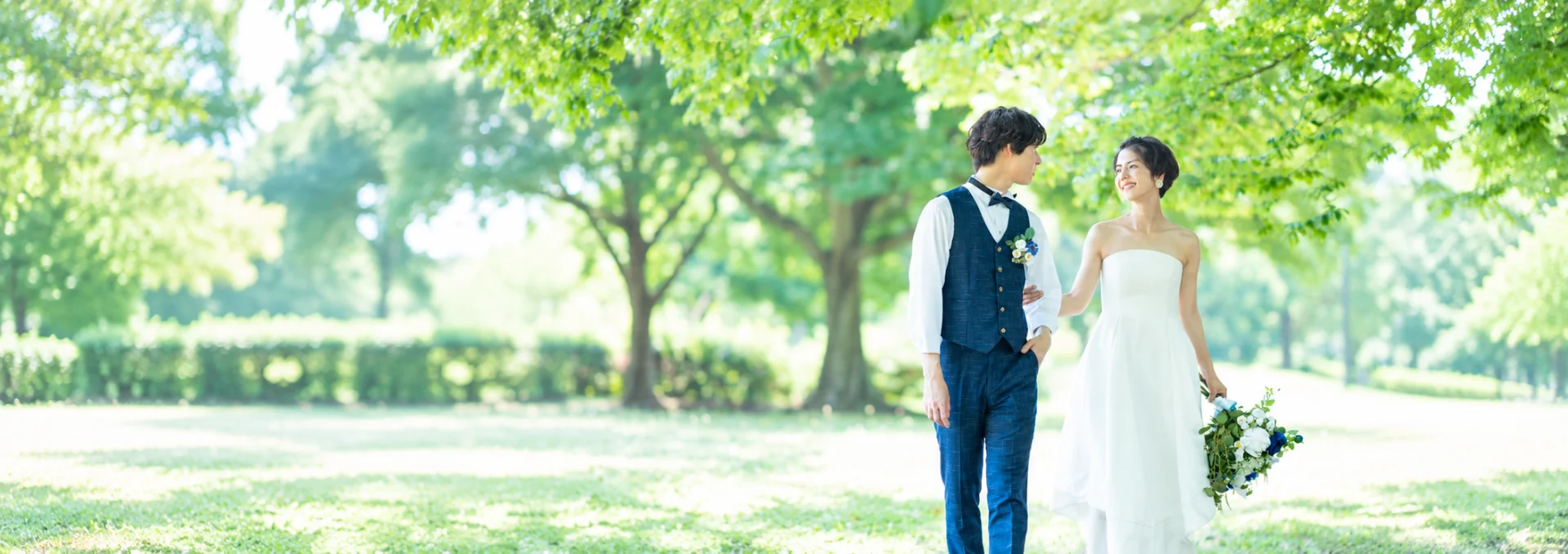 木漏れ日の公園を並んで歩き、見つめ合うウェディング姿の男女